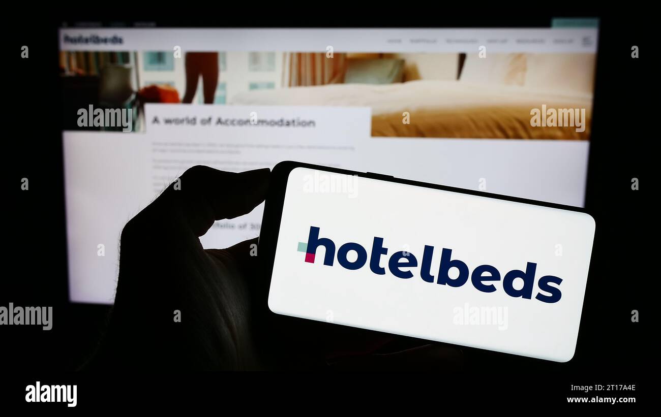 Person, die ein Mobiltelefon mit dem Logo des Reisetechnologieunternehmens Hotelbeds Group S.L.U. vor der Webseite hält. Schwerpunkt auf dem Display des Telefons. Stockfoto