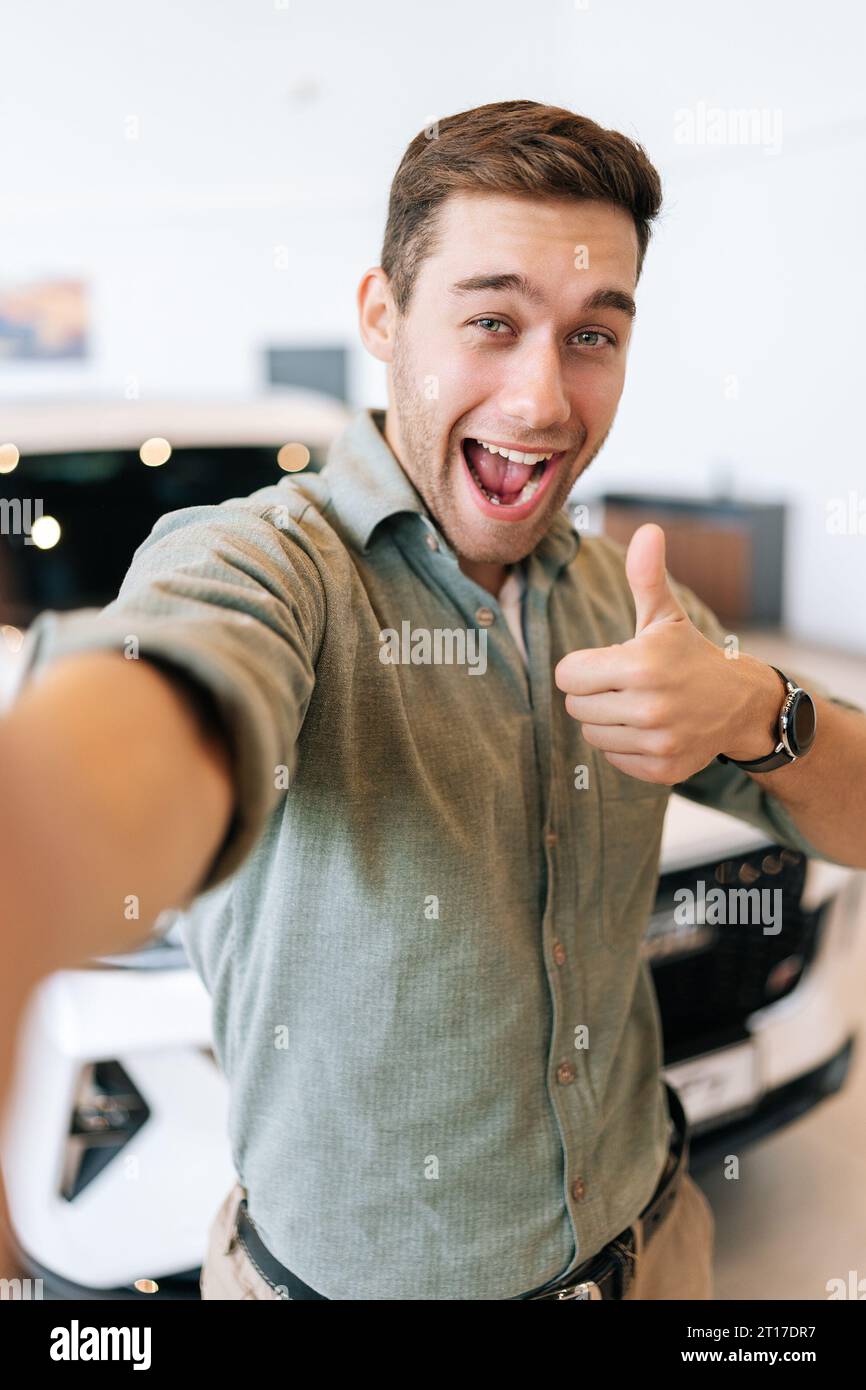 Vertikales POV-Bild eines fröhlichen Käufers, der ein Selfie mit einem Schild auf dem Smartphone vom Händlerbetrieb macht, nachdem er ein neues Auto gekauft hat. Stockfoto