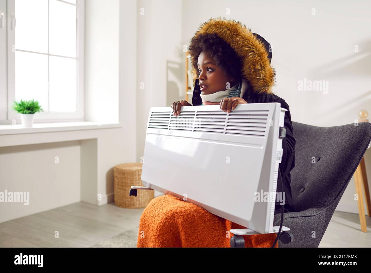 Junge hübsche afroamerikanische Frau sitzt im Sessel in warmer Jacke und Schal mit Heizung in den Händen im Wohnzimmer, friert vor Kälte, Seite Stockfoto
