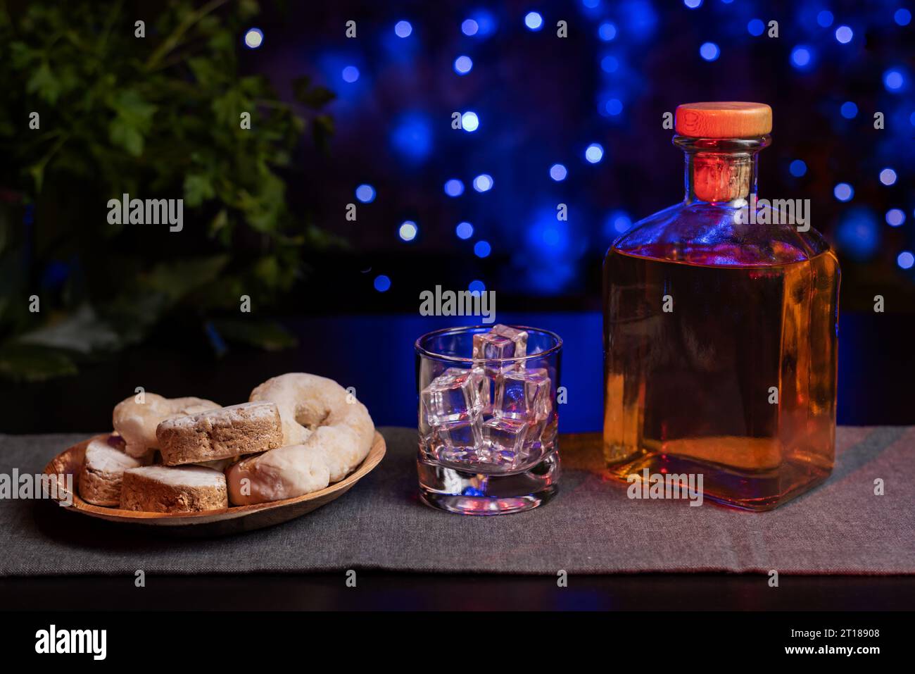 Shortbread, Wein Donuts und Blätterteiggebäck auf einem Holzteller und neben einem Glas mit Eis und einer Flasche Alkohol, mit einem Hintergrund von blauen Lichtern mit Stockfoto