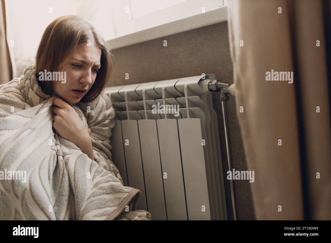 Eine leidende Frau im Karo sitzt an der Heizbatterie, wärmt sich im Haus auf und friert ein. Stockfoto