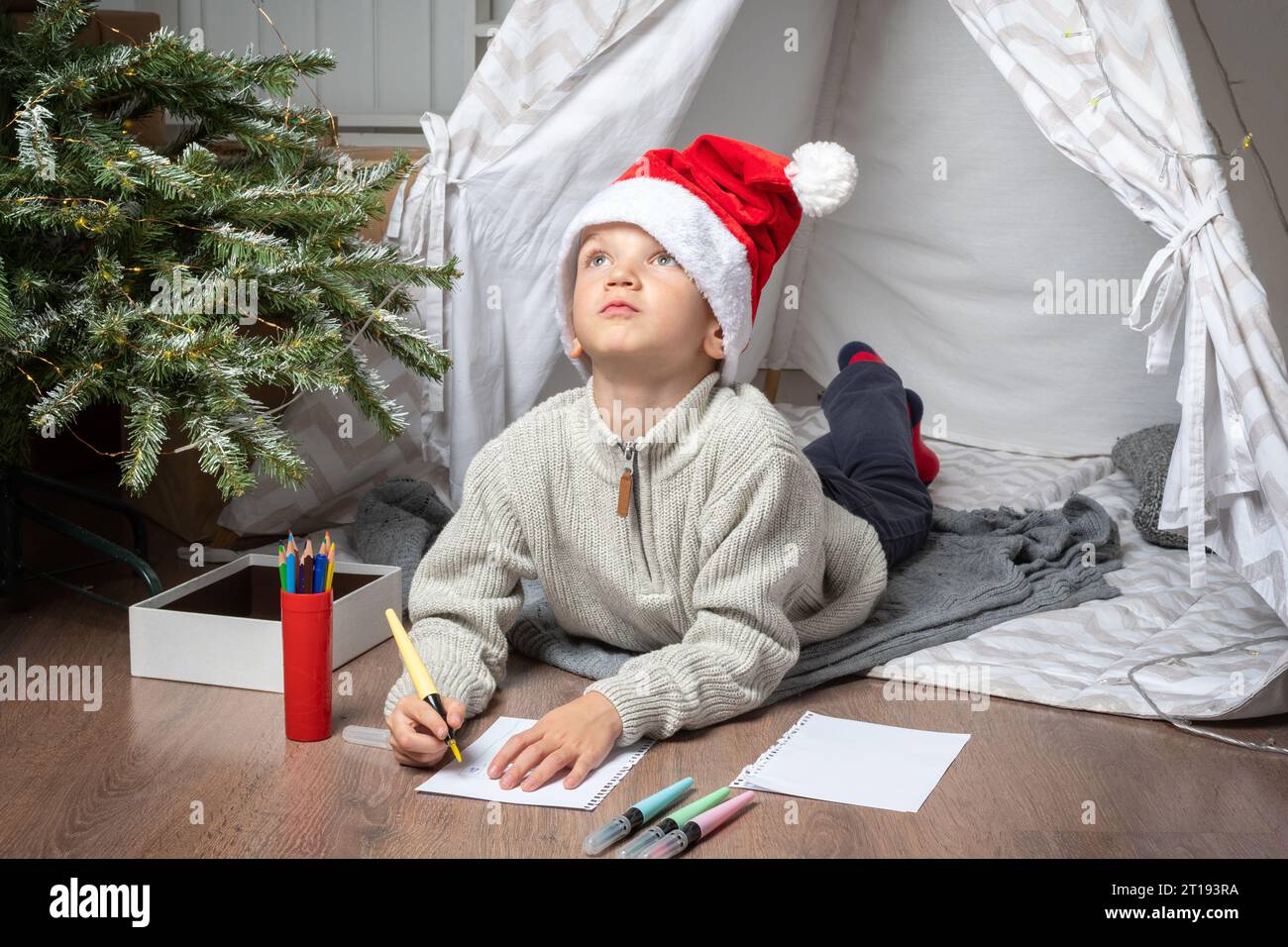 Wunschliste für Weihnachtswunder. Der lächelnde Junge im Weihnachtsmann schreibt Briefe für Geschenke an den Weihnachtsmann. Das Kind hofft auf ein Neujahrswunder Stockfoto