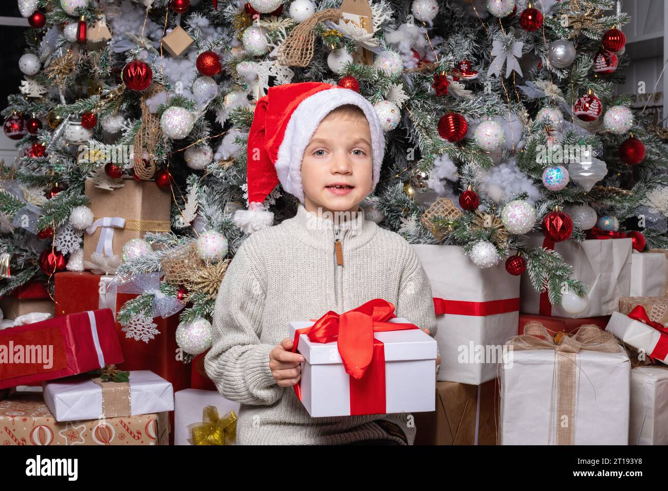 Süßes glückliches Kind mit weihnachtsmannmütze mit einem Überraschungsgeschenk auf dem Hintergrund eines dekorierten Weihnachtsbaums. Zufriedener Junge bekam ein Weihnachten oder Neujahr Stockfoto