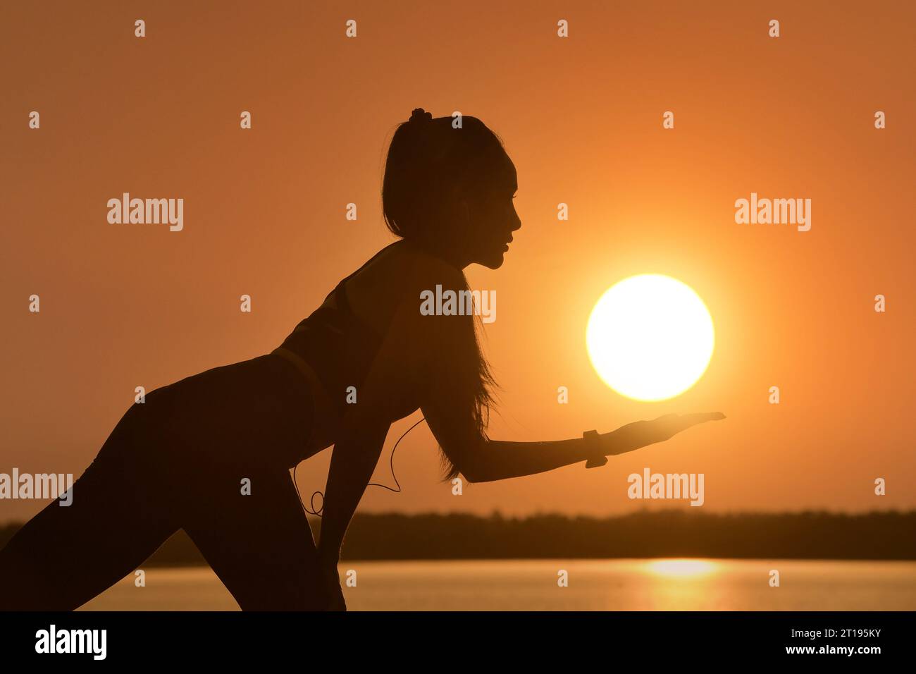 Silhouette einer Frau, die Musik hört, während sie beim Sonnenuntergang an einem See Yoga macht, Thailand Stockfoto
