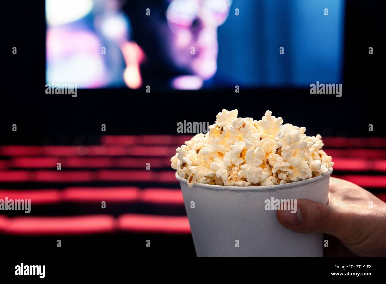 Filme und Popcorn. Ein Mann hält Popcorn-Box im Kino. Action, Thriller oder SciFi-Unterhaltung auf dem Bildschirm. Rote Sitze im dunklen Theater. Salziger Snack. Stockfoto