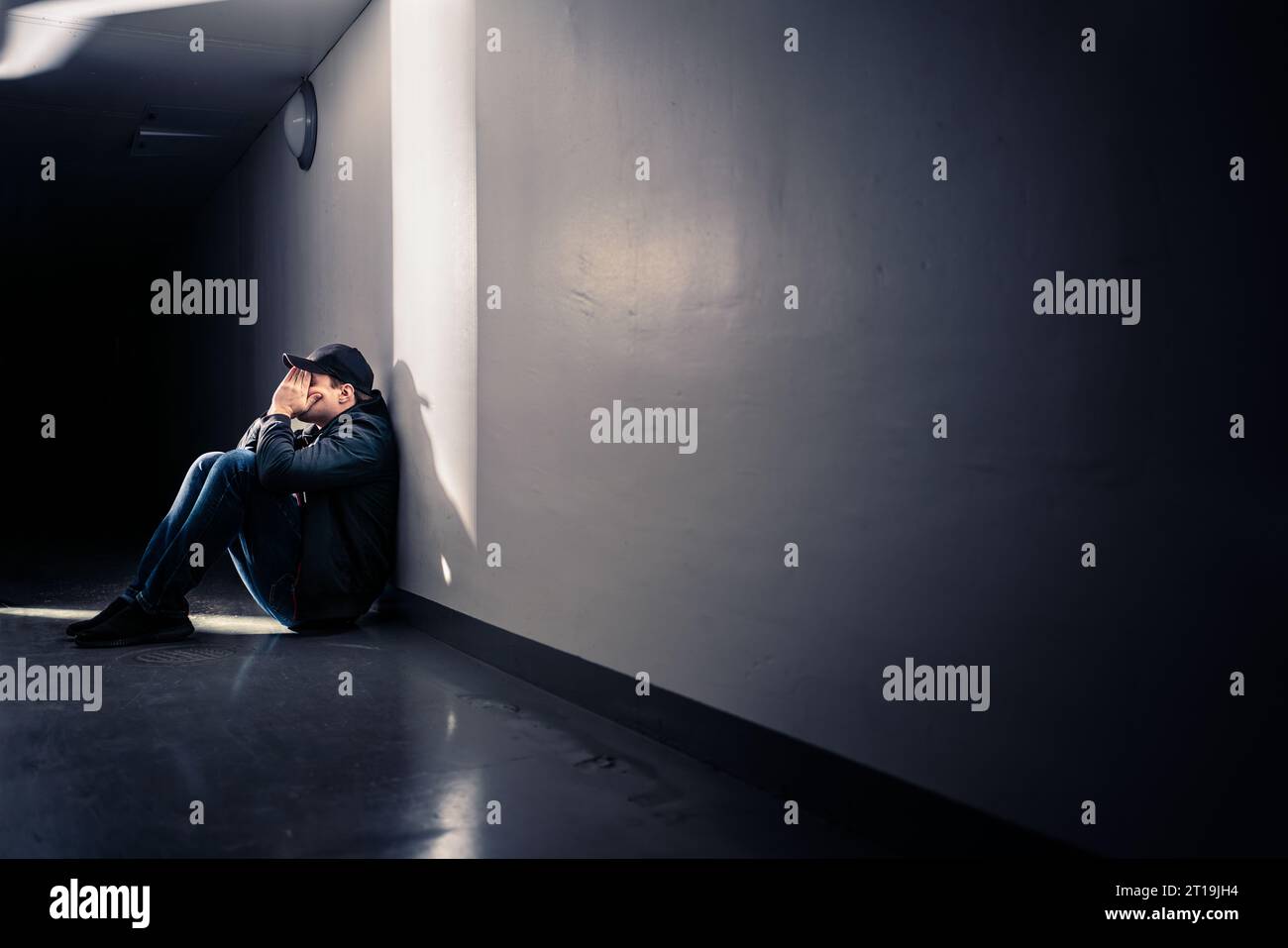 Trauriger Mann mit Trauma, der auf dem Boden sitzt. Schande, Schuld oder Trauer. Ein verzweifelter Kerl weint im dunklen Korridor. Opfer von Einsamkeit oder Diskriminierung. Stockfoto