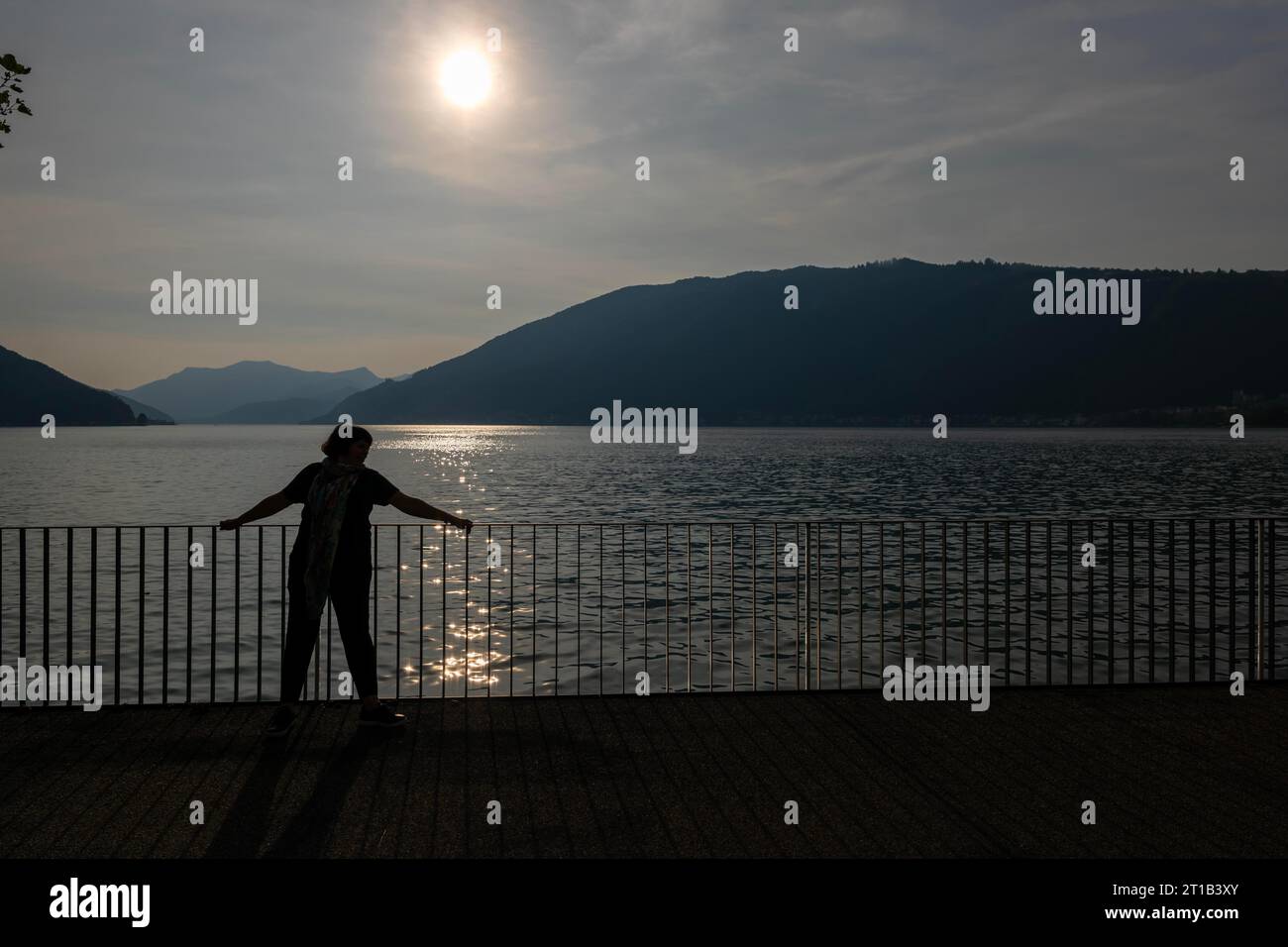 Frau am Ufer mit Geländer zum Luganersee mit Berg an einem sonnigen Sommertag in Bissone, Tessin, Schweiz Stockfoto