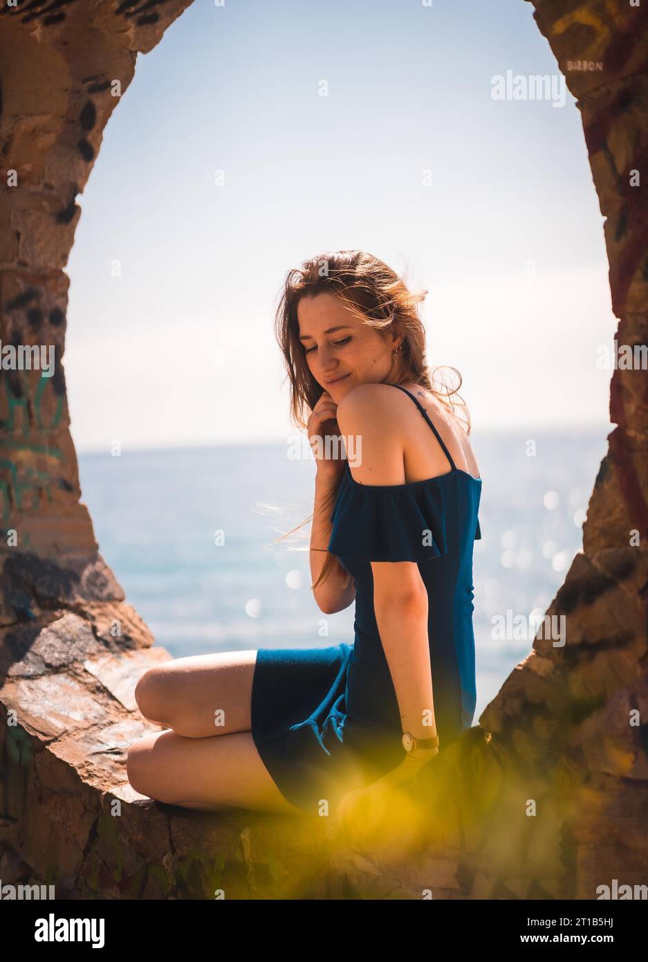 Lebensstil einer jungen, hübschen, rothaarigen Kaukasierin, die in einem blauen Kleid am Meer sitzt, in einem runden Steinfenster Stockfoto