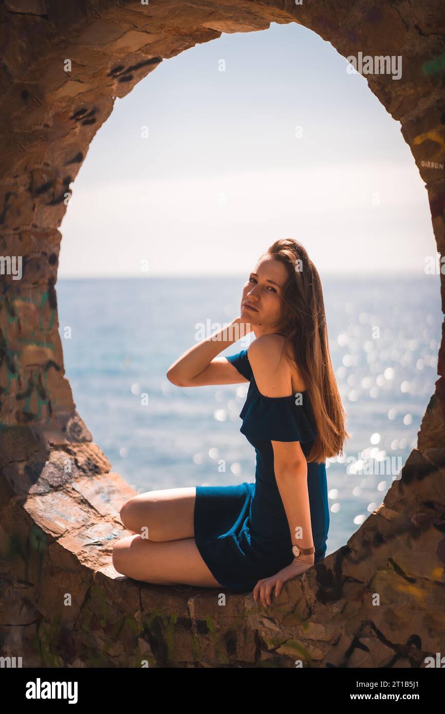 Lebensstil einer jungen, hübschen, rothaarigen Kaukasierin, die in einem blauen Kleid am Meer sitzt, in einem Steinfenster Stockfoto
