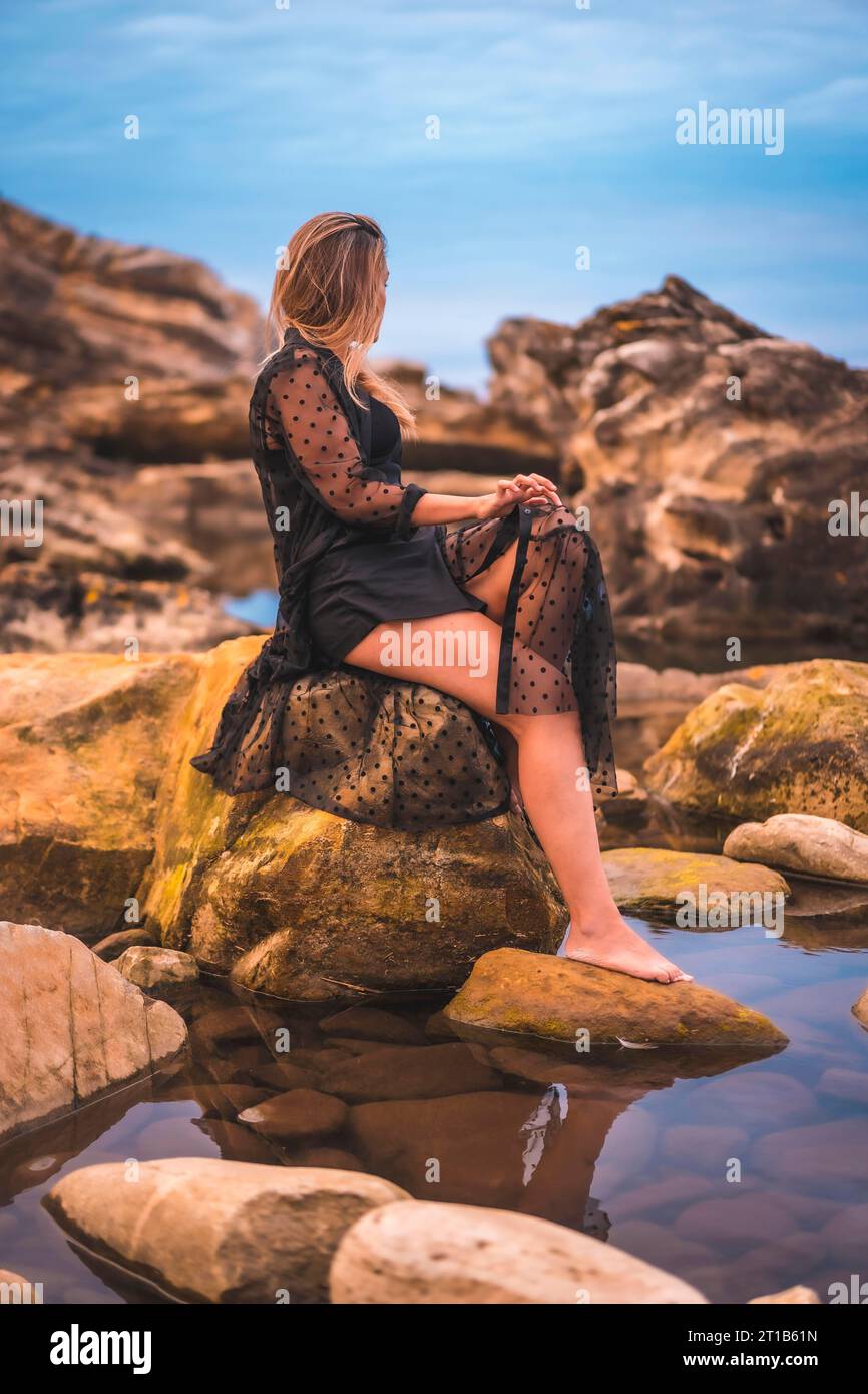 Sommer-Lifestyle mit einer jungen brünetten Kaukasierin in einem langen schwarzen transparenten Kleid auf einigen Felsen in der Nähe des Meeres an einem Sommernachmittag. Sitzen Sie auf Stockfoto