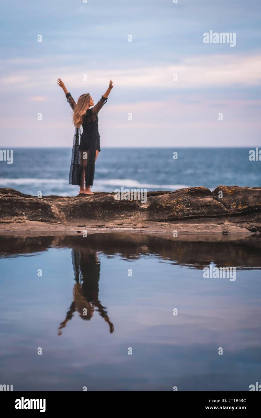 Sommer-Lifestyle mit einer jungen brünetten Kaukasierin in einem langen schwarzen transparenten Kleid auf einigen Felsen in der Nähe des Meeres an einem Sommernachmittag. Anheben Stockfoto