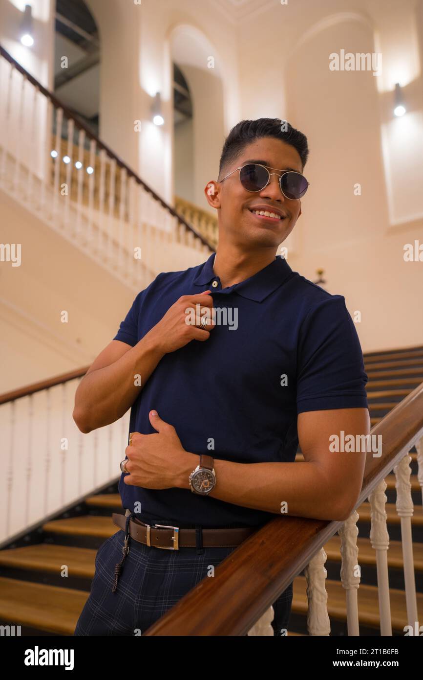 Fashion Lifestyle, Porträt eines jungen Latinos in einem schönen alten Gebäude. Blaues Poloshirt und karierte Hose. Porträt an der Treppe Stockfoto