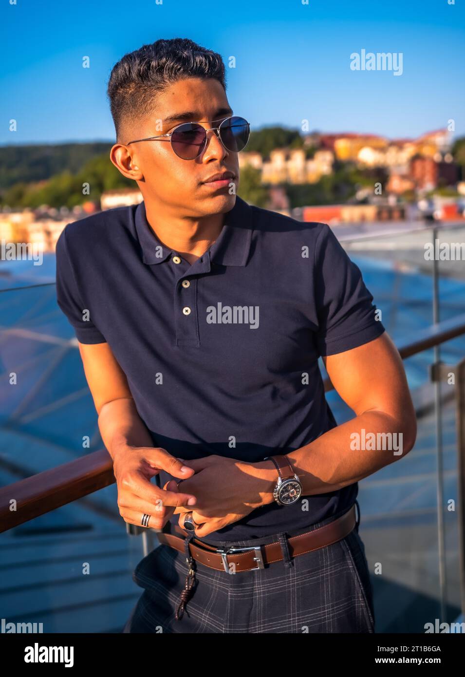 Fashion Lifestyle, Porträt eines jungen Latinos mit der Stadt San Sebastian im Hintergrund, Gipuzkoa. Blaues Poloshirt und karierte Hose, nachdenklich Stockfoto