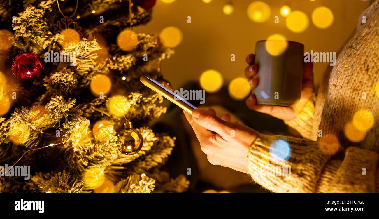Seitenansicht von Smartphone und Tasse in weiblichen Händen vor dem Weihnachtsbaum. Stockfoto