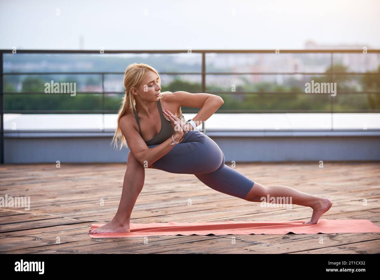 Junge Frau mit blonden Haaren und schlankem Körper macht Yoga-Übungen auf der Yoga-Matte auf der Holzterrasse bei Sonnenuntergang. Stockfoto