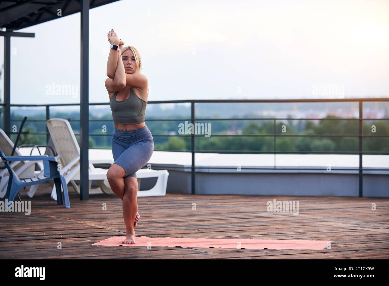 Junge Frau mit blonden Haaren und schlankem Körper macht Yoga-Übungen auf der Yoga-Matte auf der Holzterrasse bei Sonnenuntergang. Stockfoto