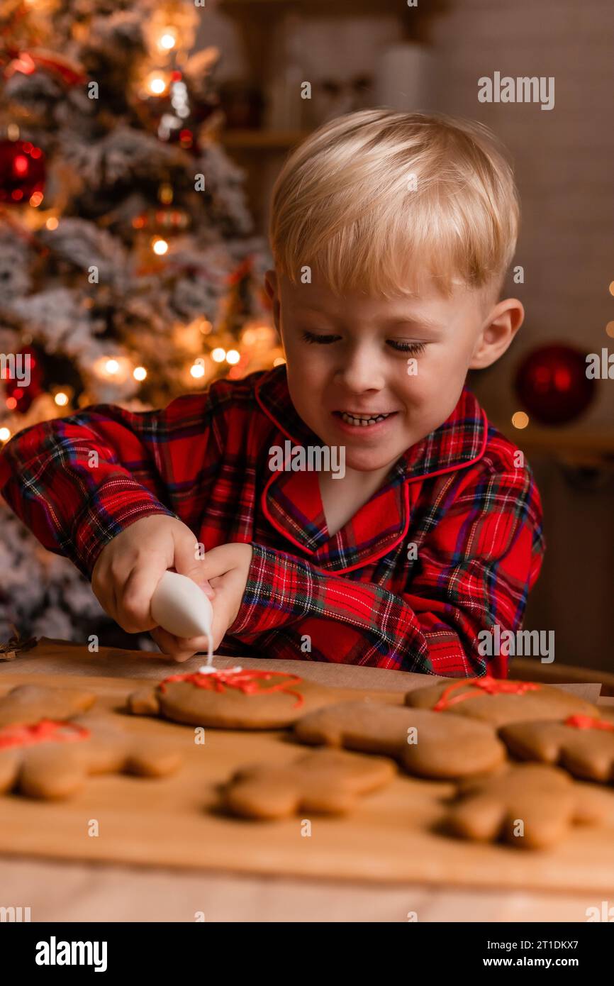 Der blonde Junge im roten karierten Pyjama schmückt Weihnachtskekse mit Glasur in der Küche. Stockfoto