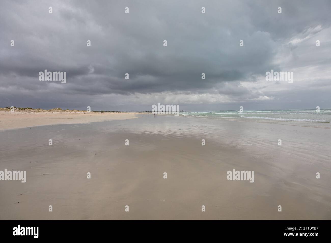 Ein riesiger Strand ist von grauen, stürmischen Wolken bedeckt, und ihre Reflexion glitzert auf dem Wasser, während entfernte Figuren entlang der Küste spazieren Stockfoto