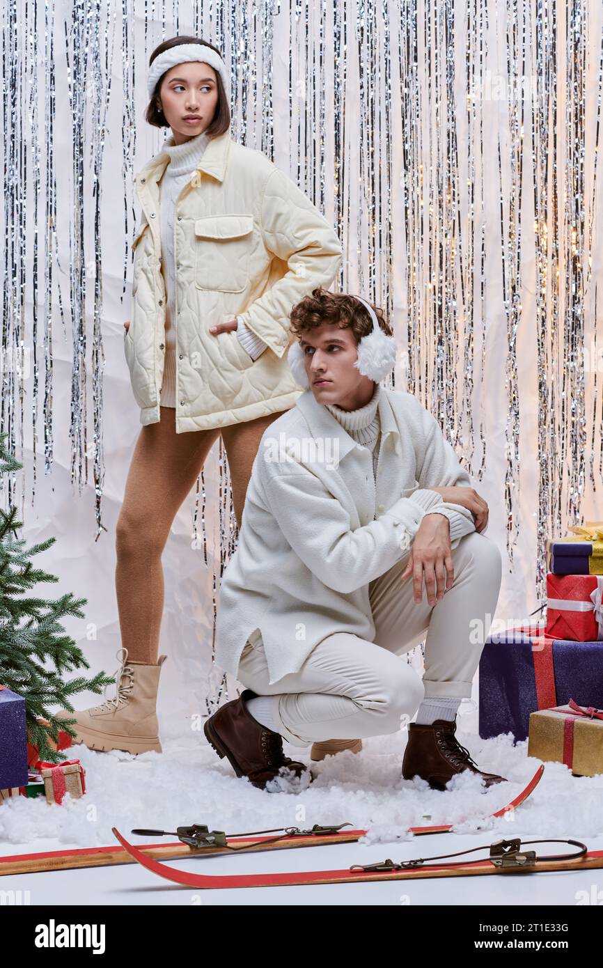 Interrassische Modelle in warmen Outfits, die neben dem weihnachtsbaum posieren, Geschenke und Silberdekor im Studio Stockfoto