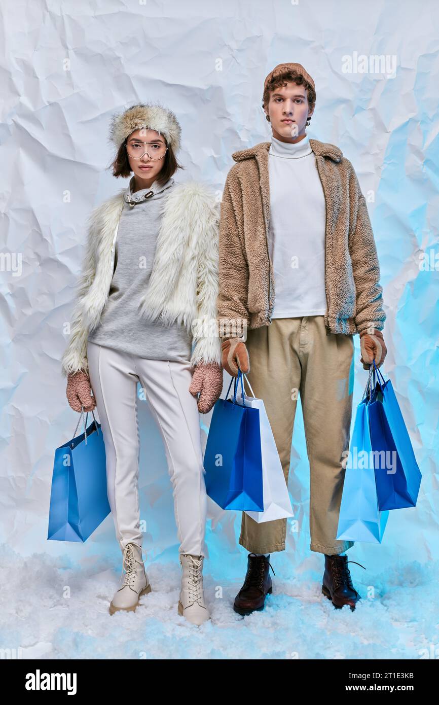 Trendige Interracial Models in Winterkleidung mit blauen Einkaufstaschen und Blick auf die Kamera im verschneiten Studio Stockfoto