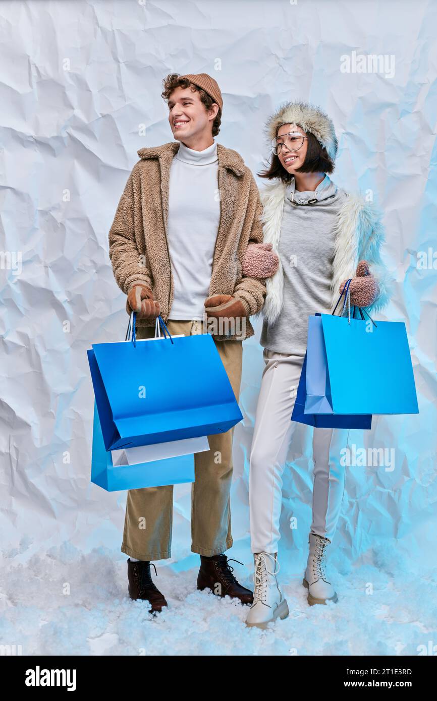 Glückliches interrassisches Paar in Winterkleidung mit blauen Einkaufstaschen im verschneiten Studio, saisonale Einkäufe Stockfoto