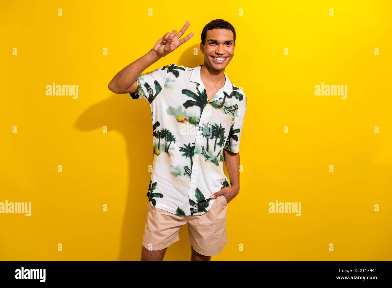 Porträt des angenehmen funky Guy mit Ring in der Nase tragen bedruckte Kleidung zeigt V-Zeichen Hand in der Tasche isoliert auf gelbem farbigen Hintergrund Stockfoto