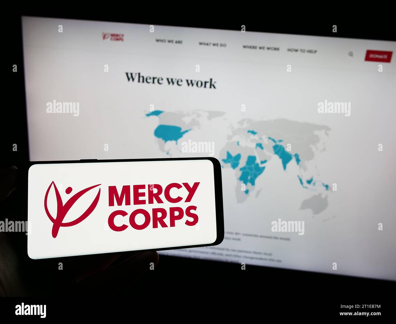 Person mit Handy mit Logo der US-Hilfsorganisation Mercy Corps vor der Webseite. Schwerpunkt auf dem Display des Telefons. Stockfoto