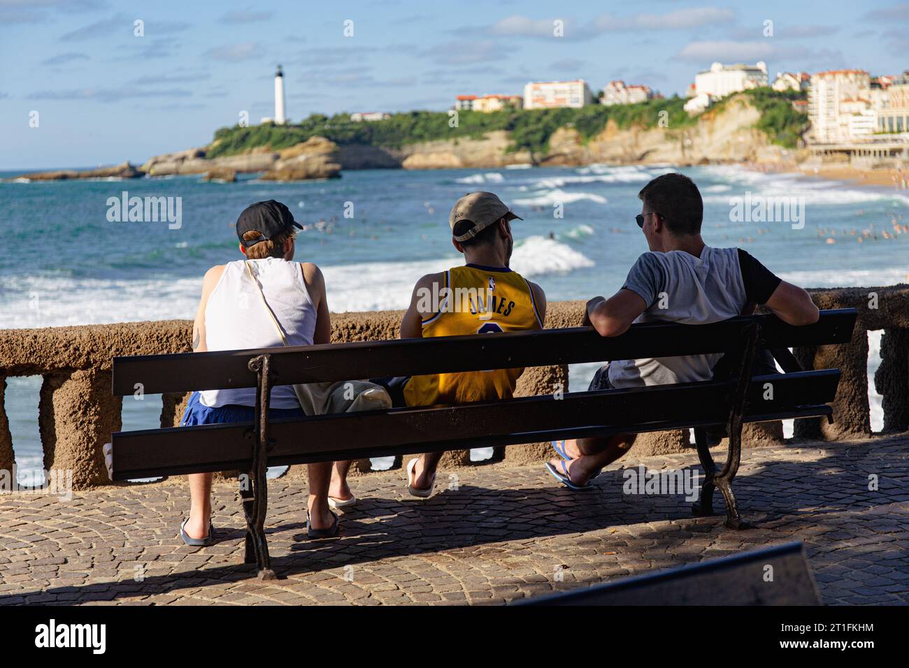 Drei Männer sitzen auf einer Bank und starren auf den Strand in Biarritz. Leuchtturm weit weg im Hintergrund. Stockfoto