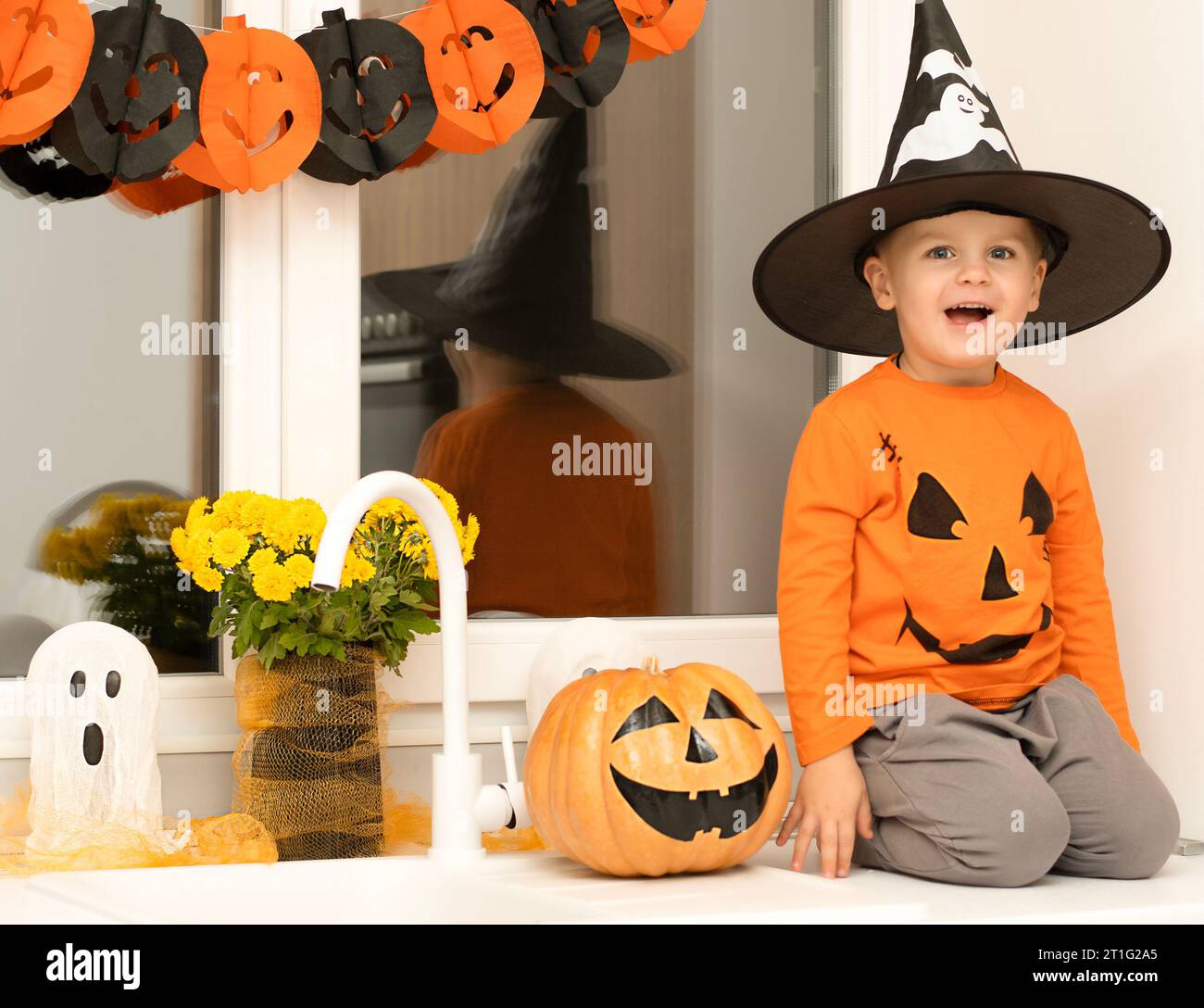 Halloween-Konzept. Ein kleiner, fröhlicher, gutaussehender Junge mit Zaubererhut und orangefarbenem Pullover sitzt auf einem Tisch in der Küche vor dem Hintergrund eines Stockfoto