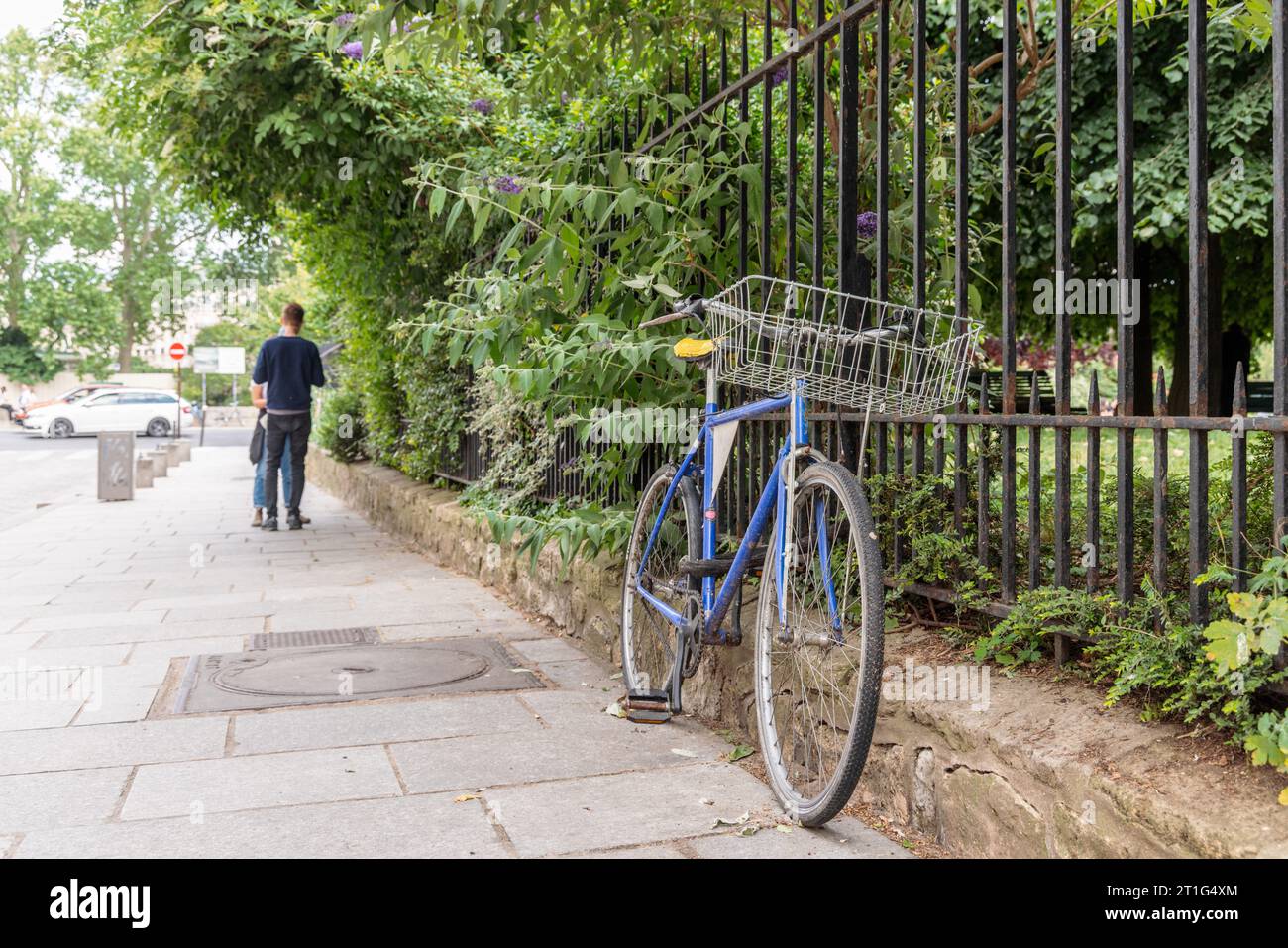 Blaues Fahrrad ist in Paris, Frankreich, an Geländer gekettet. Im unscharfen Hintergrund unterhalten sich zwei Personen. Autos parkten in der Ferne. Stockfoto