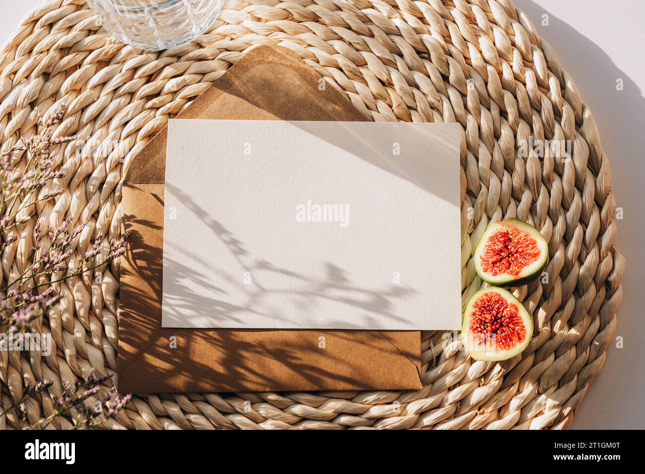 Umschlag mit leerer Karte auf einer Korbmatte mit Feigenfrüchten und Blumen Schatten. Draufsicht, flach, Modell. Stockfoto