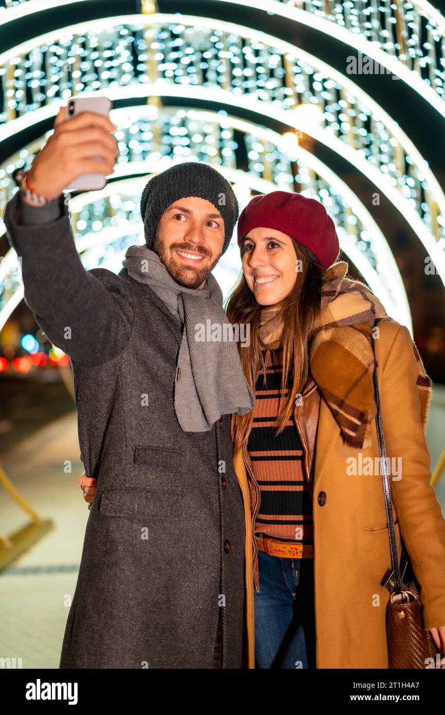 Ein europäisches Paar, das die Weihnachtslichter der Stadt genießt, macht ein Selfie mit dem Handy für soziale Netzwerke und Lifestyle Stockfoto