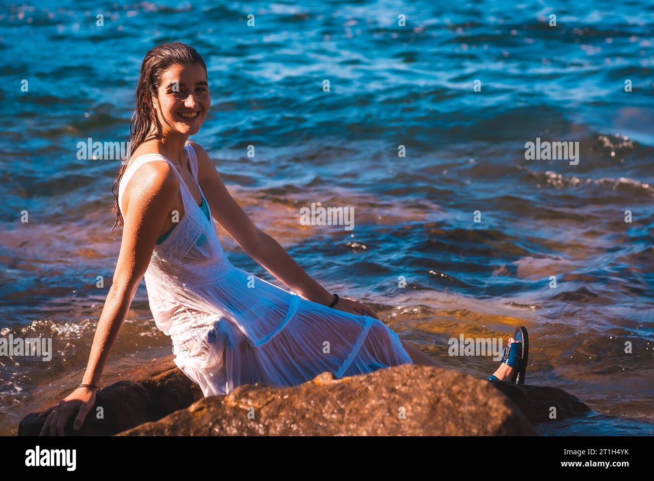 Ein junges kaukasisches Mädchen, das in einem nassen weißen Kleid lächelt Auf einem Felsen sitzend Stockfoto