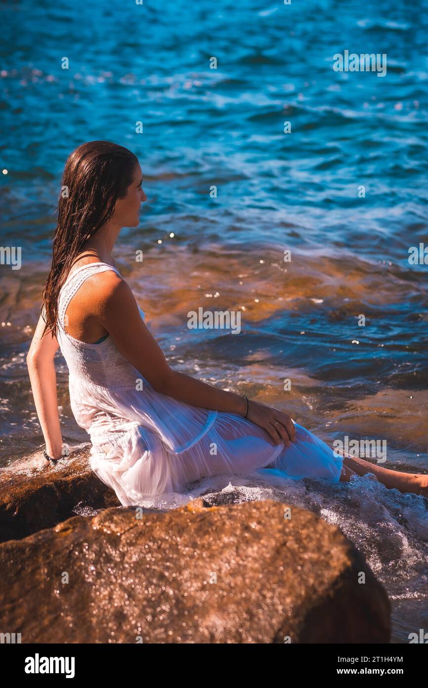 Eine junge kaukasische Frau in einem nassen weißen Kleid sitzend Auf einem Felsen Stockfoto