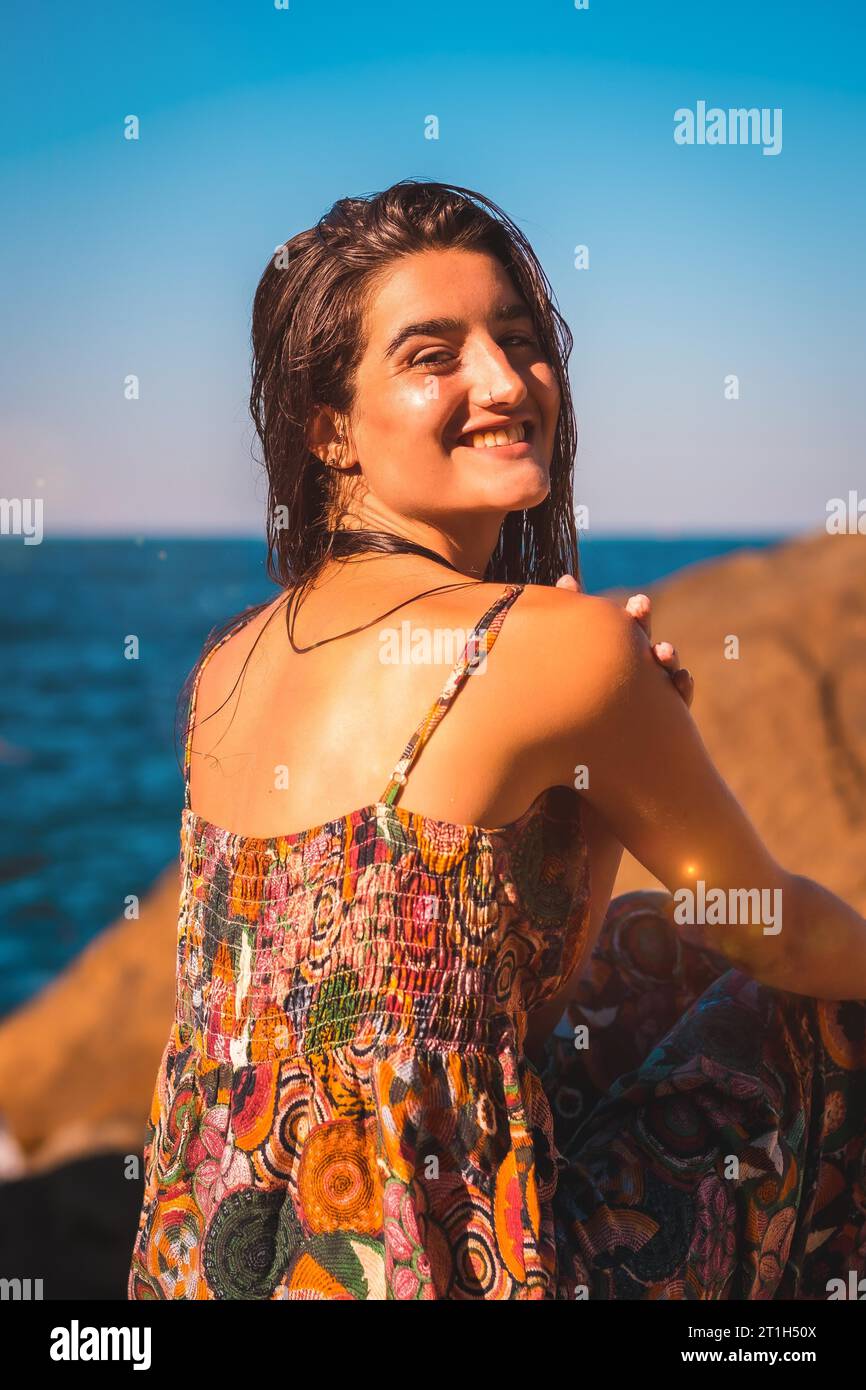 Porträt einer jungen kaukasischen Frau von hinten, die mit nassen Haaren und einem Blumenkleid am Meer lächelt, Sommerleben Stockfoto