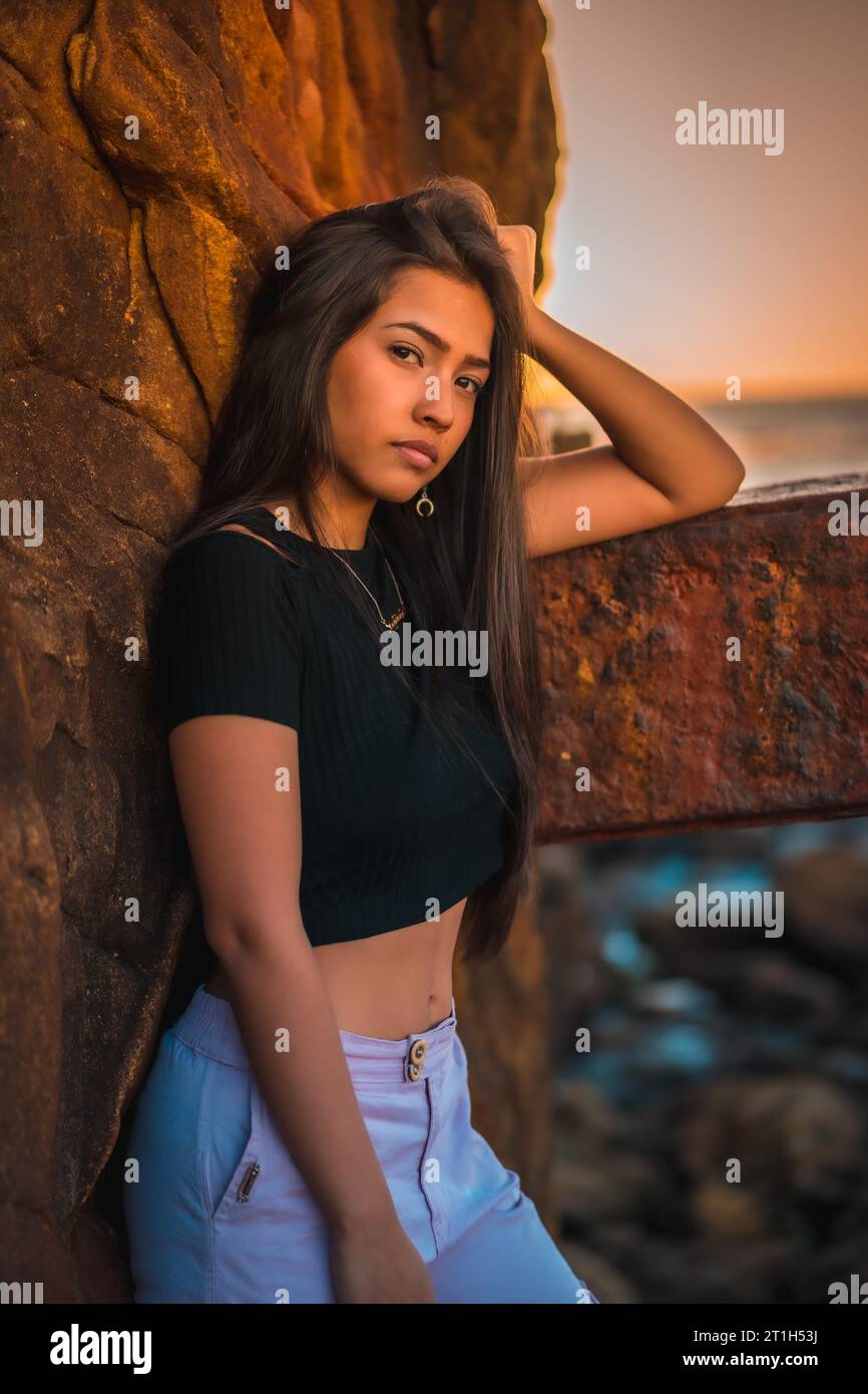 Eine junge hübsche Brünette Latina mit langen glatten Haaren in einem kurzen schwarzen T-Shirt und rosa Hose. Porträt der Frau, die sich auf eine Skulptur mit Schorf stützt Stockfoto