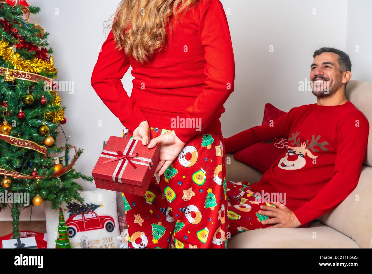 Ein junges Paar sitzt am Weihnachtsbaum, die Freundin gibt dem Freund ein Weihnachtsgeschenk Stockfoto