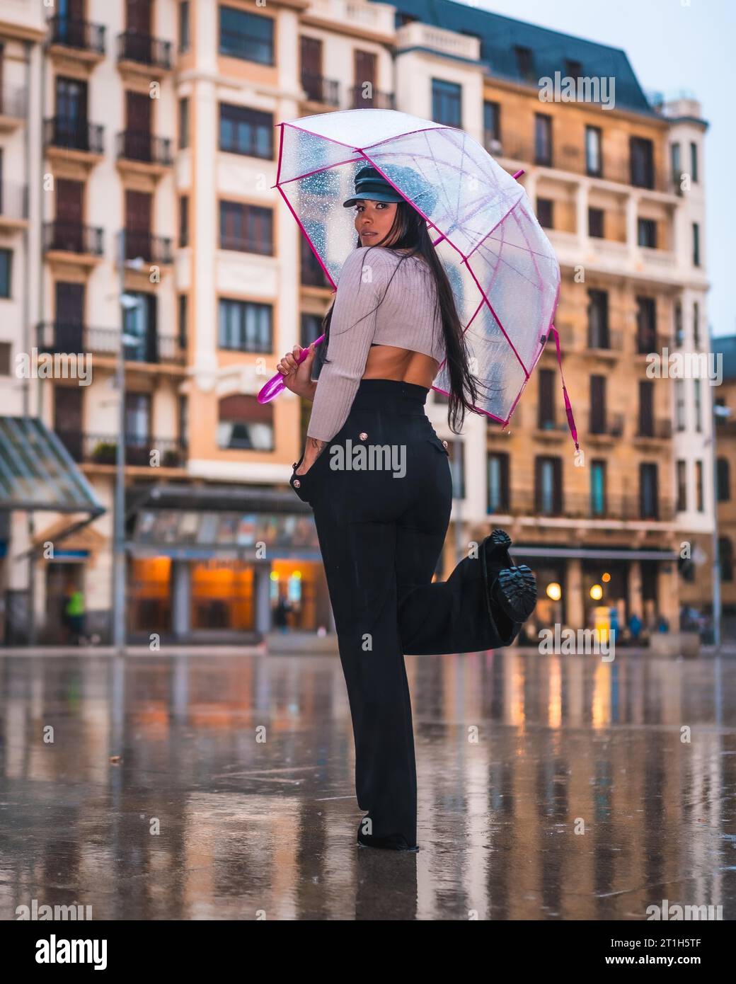 Posiert einer jungen brünetten Latina mit Ledermütze und schwarzer Hose im Herbstregen mit einem transparenten Regenschirm in der Stadt Stockfoto