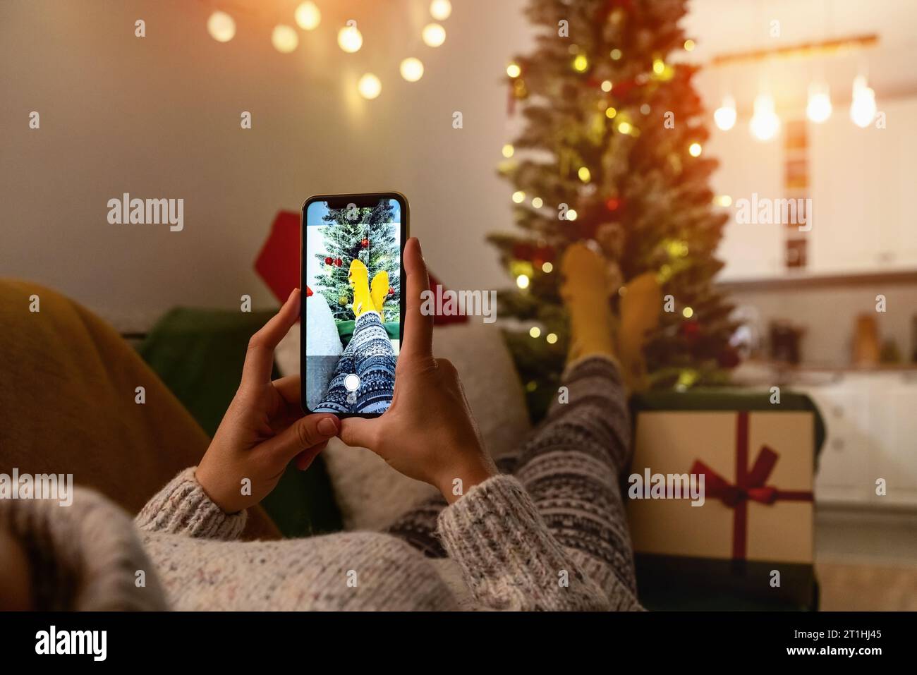 Frau, die auf dem Sofa liegt und Fotos von ihren Füßen in Socken vor dem Weihnachtsbaum macht. Fotonachrichten während der Weihnachtszeit. Stockfoto
