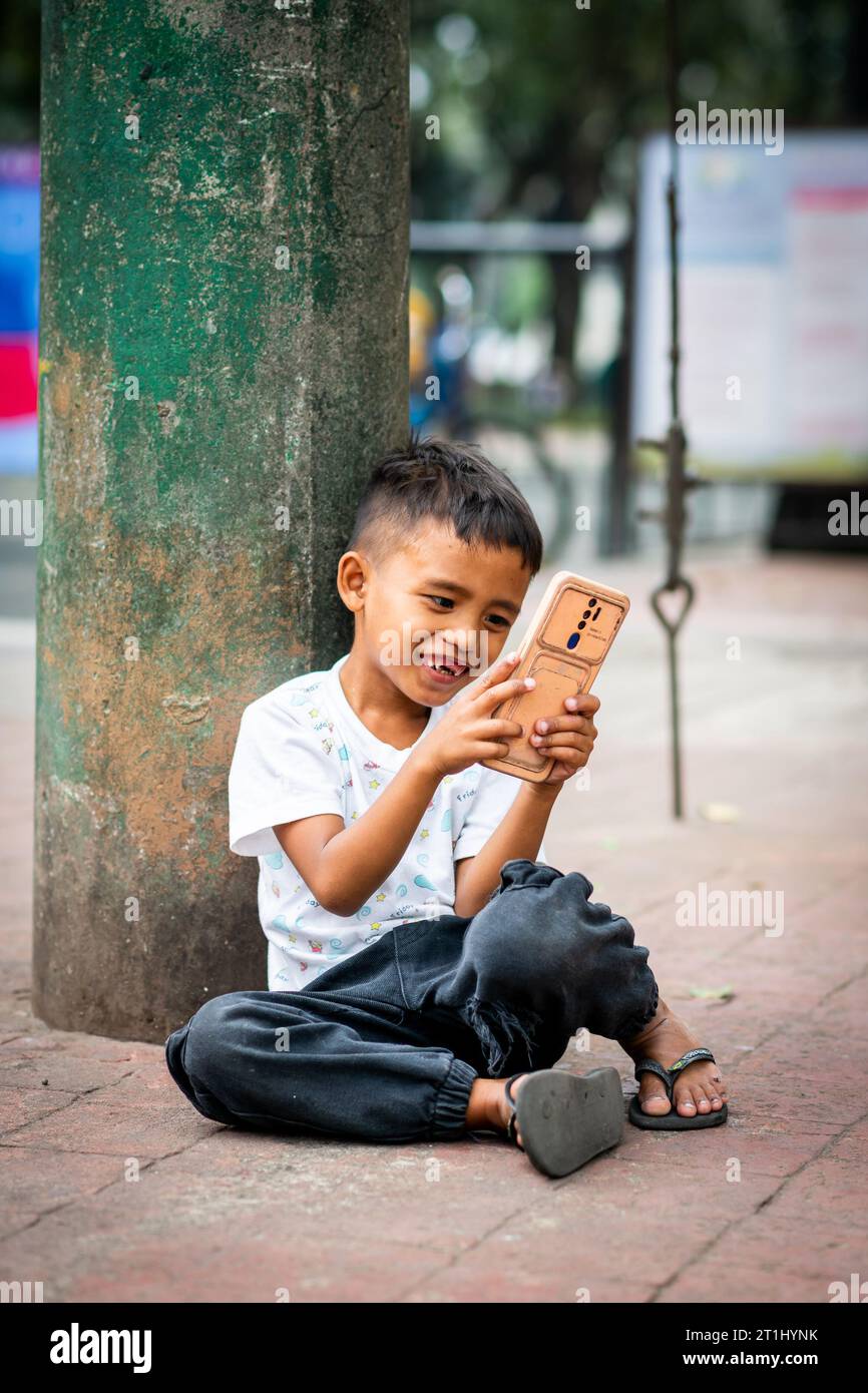 Ein junger philippinischer Junge genießt etwas auf seinem Handy, saß vor dem Rizal Park, Manila, den Philippinen. Stockfoto