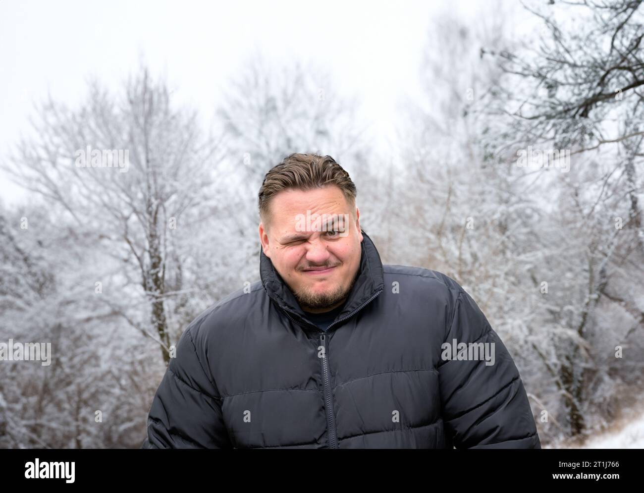 Junger Mann in einer dicken Winterjacke, der mit geschlossenem Auge schelmisch in die Kamera schaut Stockfoto