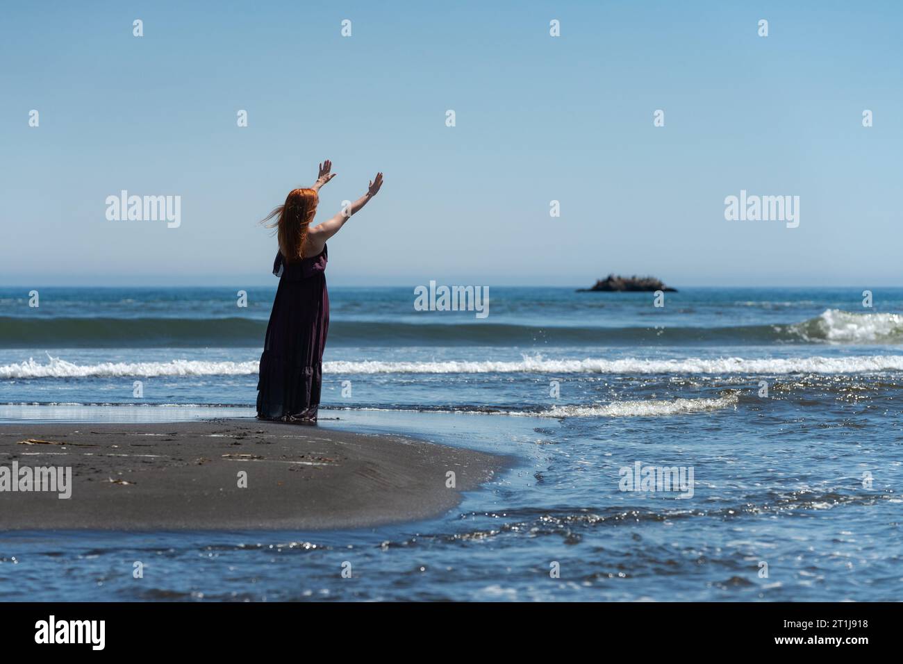 Rückansicht einer glücklichen rothaarigen Frau in dunkelviolettem, langem Kleid, die am Sommersandstrand steht und die Arme zum blauen Himmel und den Meereswellen reichen. Voll Stockfoto