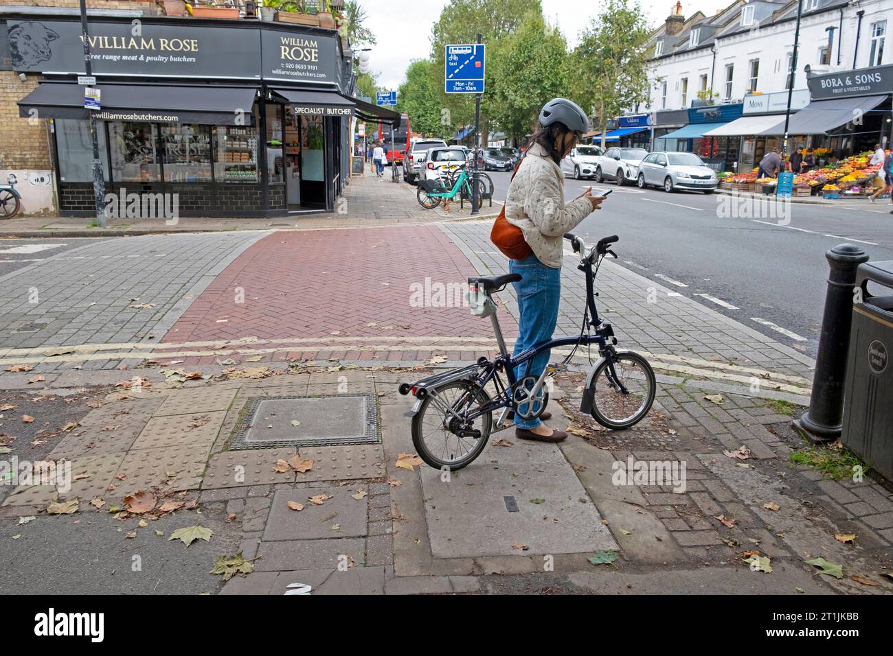 Junge Radfahrerin mit Fahrradhelm, SMS auf Handy stehend mit Brompton Fahrrad auf der Lordship Lane in East Dulwich South London SE22 Stockfoto