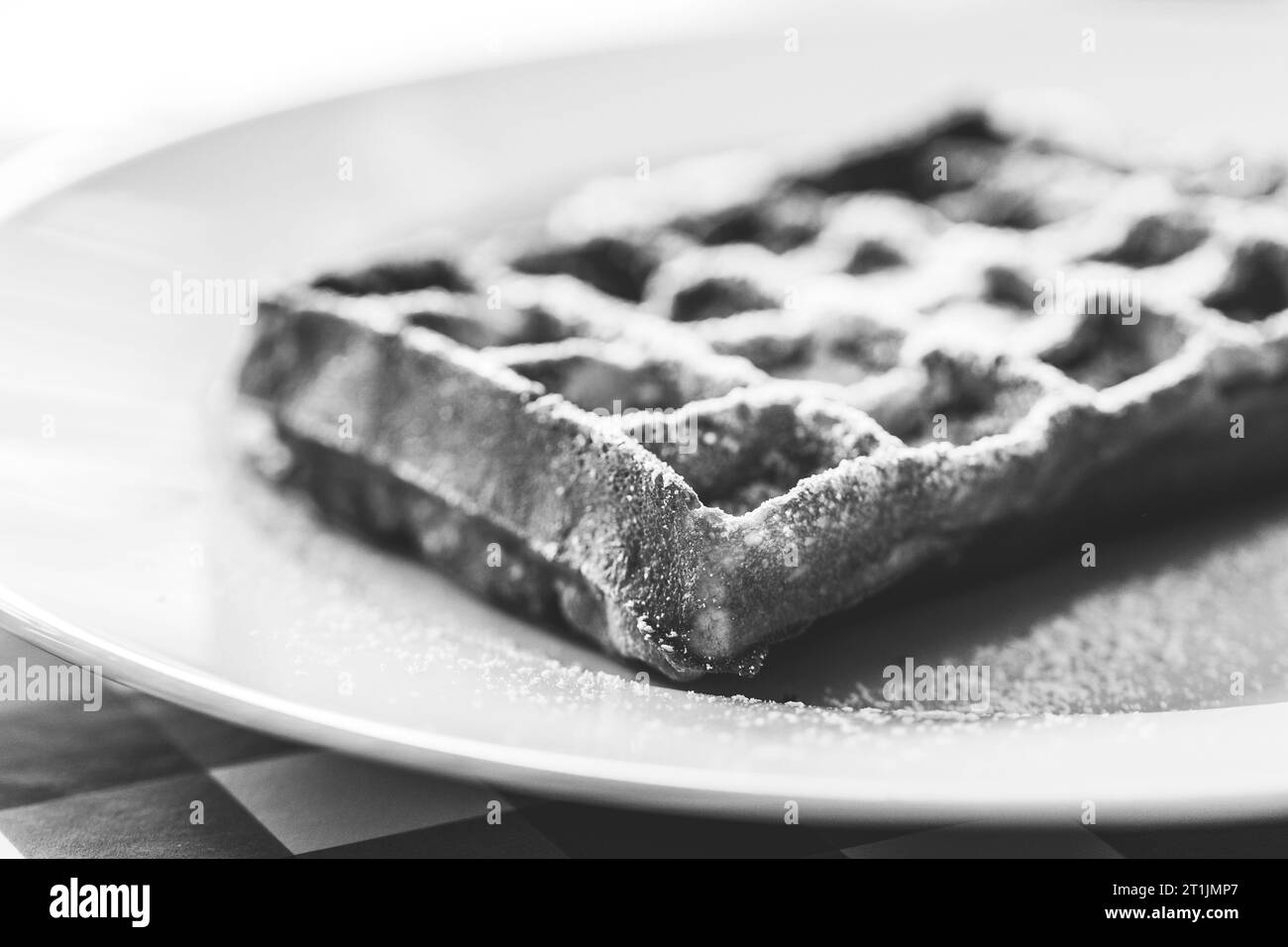 Schwarz-weißes Porträt einer leckeren belgischen Waffel mit Puderzucker als Belag, die auf einem weißen Teller auf einem roten Tischtuch liegt Stockfoto
