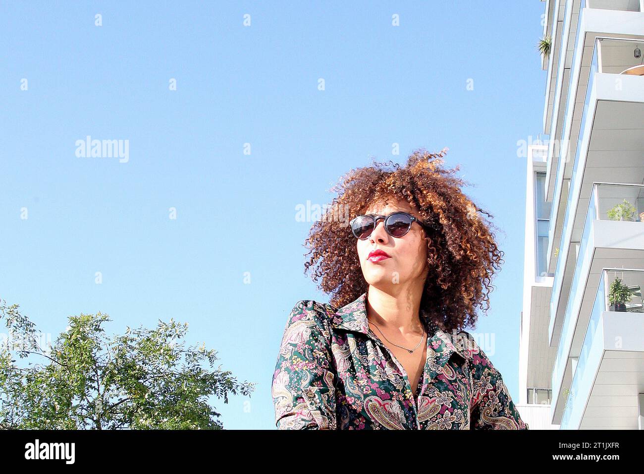 KOPFPORTRÄT EINER FRAU MIT LOCKIGEM HAAR UND SONNENBRILLE AN EINEM HEISSEN SONNIGEN TAG Stockfoto