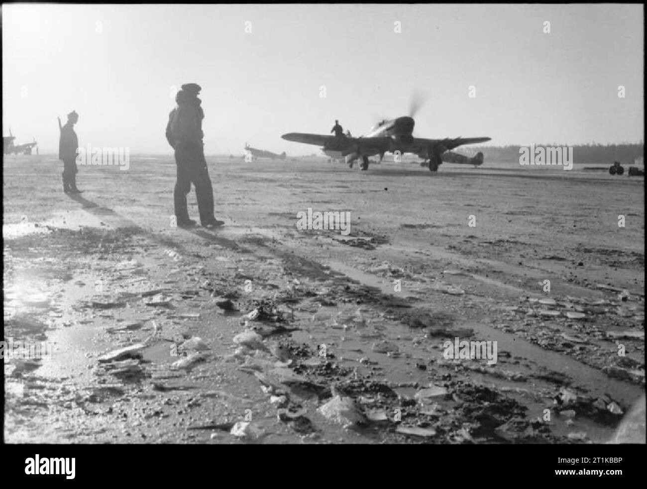 Royal Air Force - 2nd Tactical Air Force, 1943-1945. Eine Hawker Typhoon IB der Markierung Nr. 143 Flügel Taxis Vergangenheit verstreut Supermarine Spitfire Mark IXs entlang der eisbedeckten Landebahn am B78/Eindhoven, Holland, für Früh am Morgen sortie. Beachten Sie den Boden crewman sitzen auf Flügel der Typhoon der Pilot zu führen, deren Sicht nach vorn durch die Nase des Flugzeugs verdunkelt war. Stockfoto