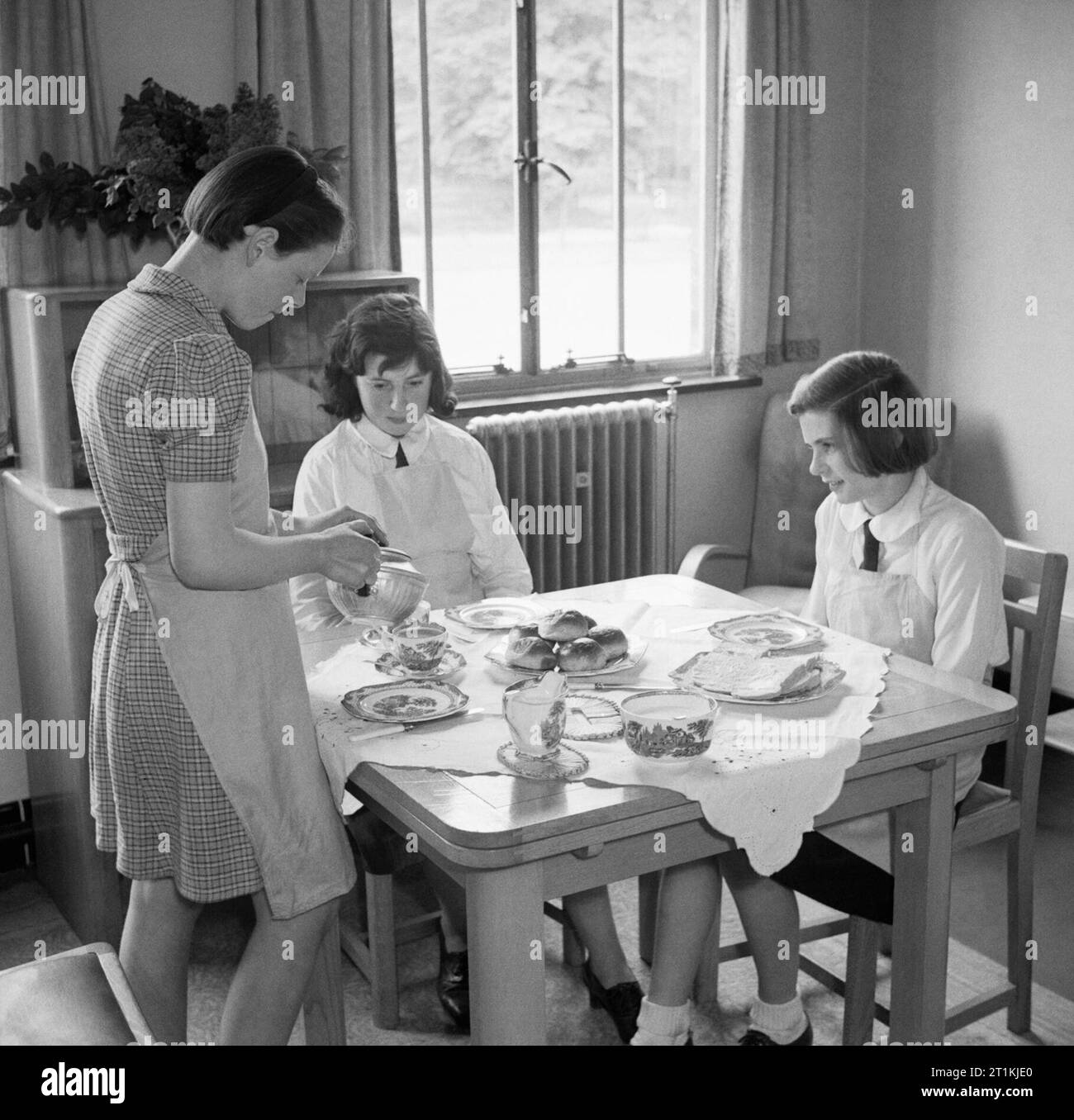 Land Schule - Alltag in Baldock County Council Schule, Baldock, Hertfordshire, England, UK, 1944 drei Mädchen in Baldock County Council Schule Kaffee nehmen im Modell flach an der Schule, die nach den ursprünglichen CAPTION,'bietet für "echte" die allgemeine Ordnung und Sauberkeit sowie die Einweisung in die Feinheiten des täglichen Lebens". Ein Mädchen steht neben den Tisch und gießt aus der Teekanne, während die anderen warten in ihren Kaffee und Brötchen zu verstauen. Stockfoto