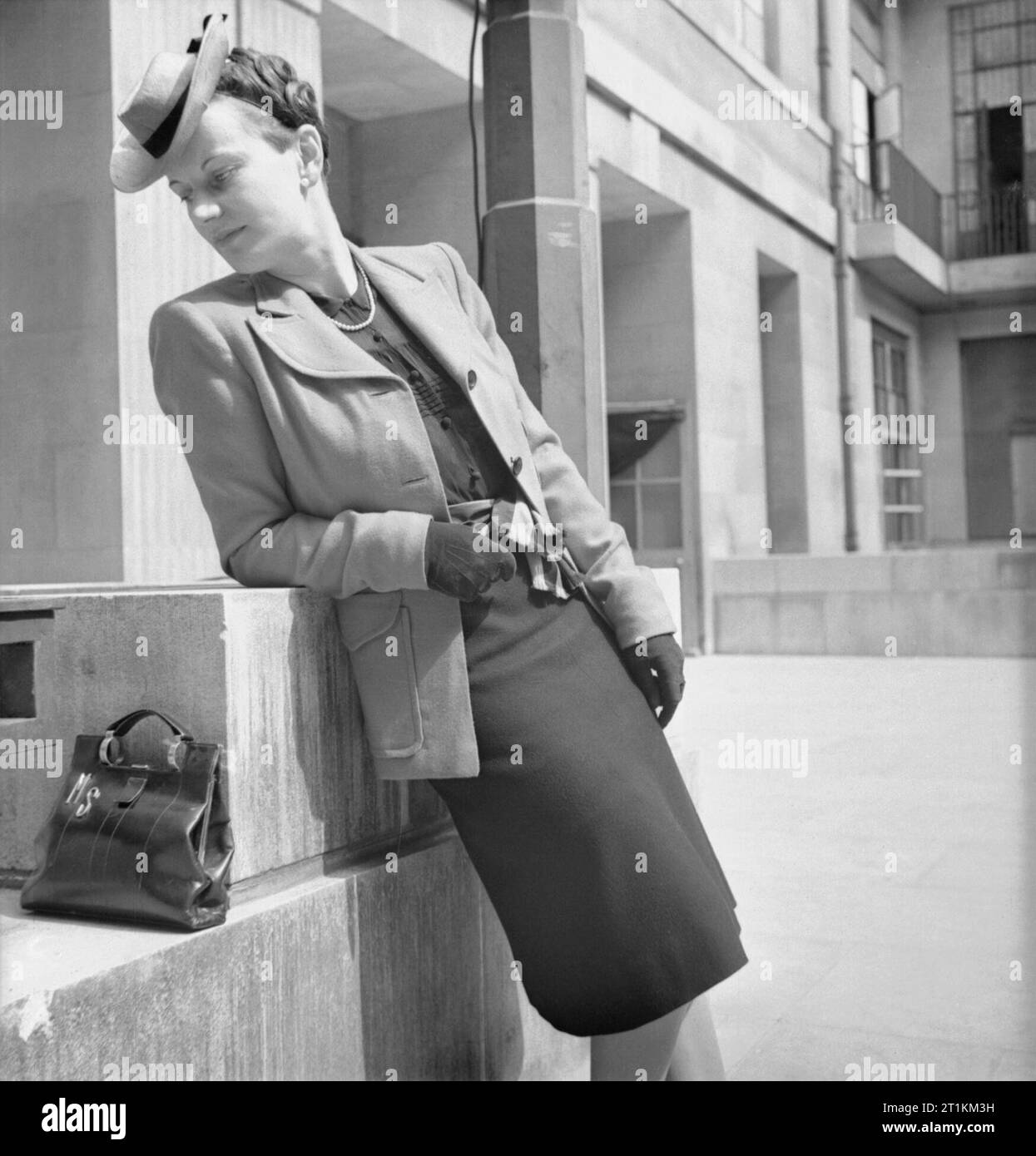 Wie eine britische Frau Kleider in Kriegszeiten - Utility Kleidung in Großbritannien, 1943 ein Modell gegen eine niedrige Mauer lehnt, ihren Kopf schräg nach unten, weg von Ihrem Outfit zu zeigen. Sie trägt die Jacke von einem cyclamen Utility Anzug, gekauft von Dickins und Jones Ltd., für 18 Coupons und 82/2 d mit einem schwarzen Rock von einer anderen Farbe. Sie ist auch mit Hut und Handschuhe und ihre Handtasche ist Aufgestützt auf der Wand neben ihr. Dieses Foto wurde am Eingang zum Senat Haus genommen, dem Sitz des Ministeriums für Informationen. Stockfoto