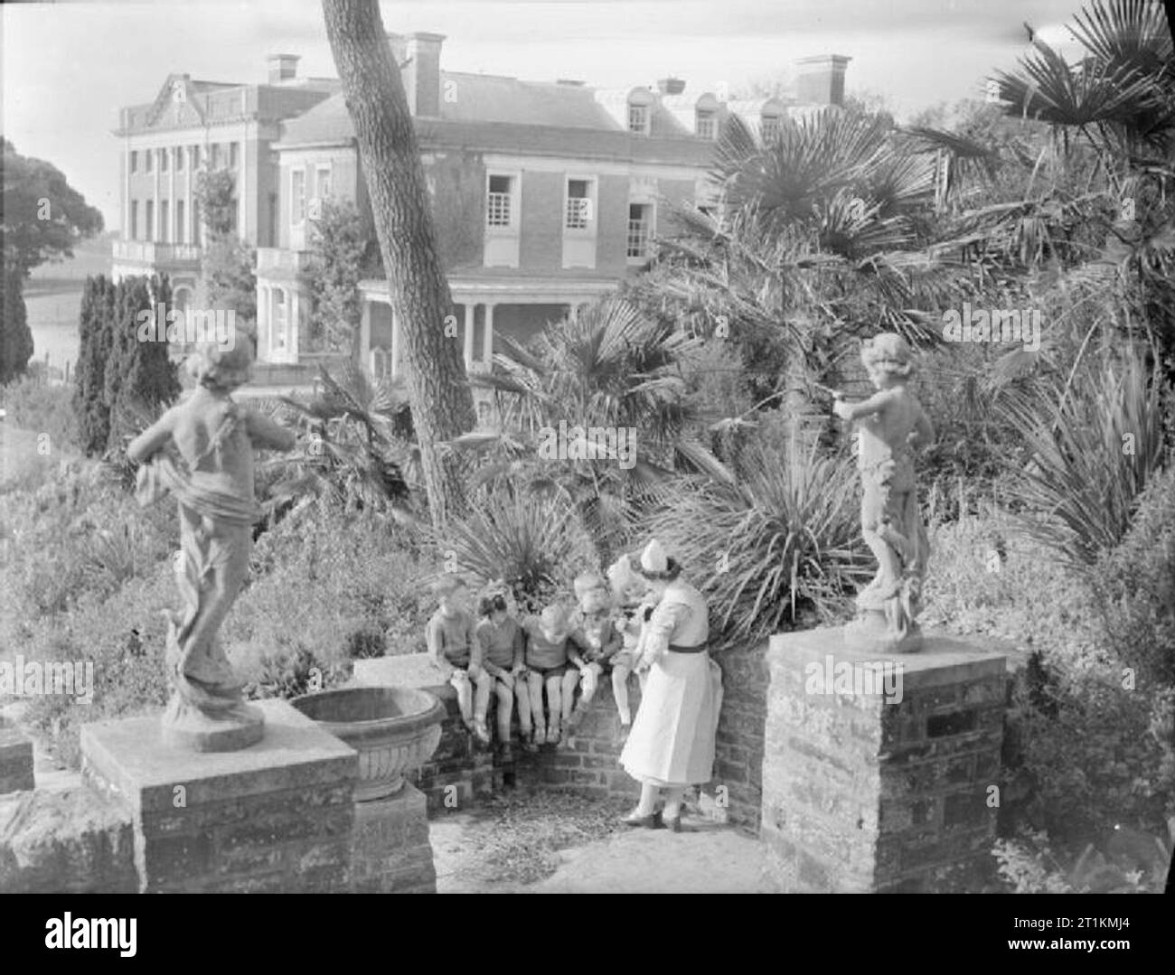 Leben an der tapley's Park Kinder Home (Chaim Weizmann Home), Instow, Devon, Oktober 1942 fünf junge Kinder sind von einer Katze, die von ihrer Schwester gehalten wird, da sie auf einem sonnigen Wand in die üppigen Gärten von tapley Park sitzen amüsiert. Es scheint, dass die Kinder auf einem Spaziergang rund um das Gelände wurden, sie sind alle mit kleinen Büscheln wilder Blumen, die Sie ausgewählt haben. Im Hintergrund die imposante Fassade des Hauses selbst gesehen werden kann. Im Vordergrund, zwei garten statuen Wache über das Verfahren. Stockfoto