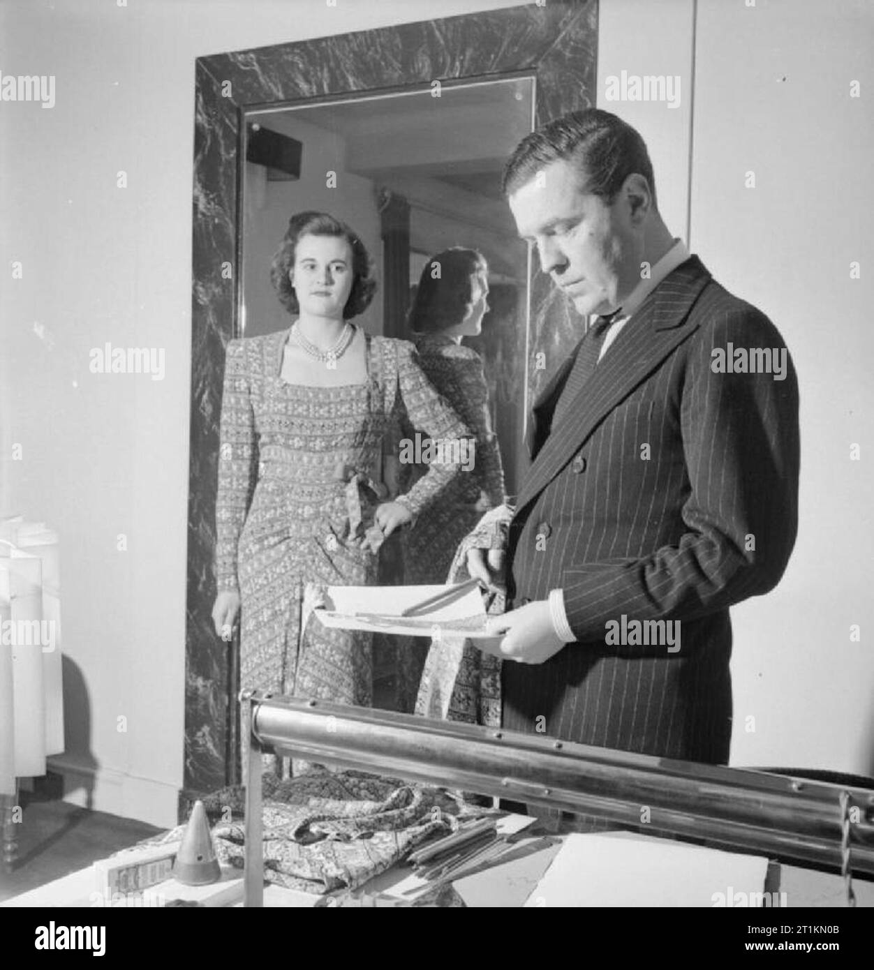 London Fashion Designer - die Arbeit der Mitglieder der Incorporated Society London Fashion Designer in Kriegszeiten, London, England, UK, 1944 In seinem Büro in London, Mode Designer Norman Hartnell vergleicht seine ursprünglichen Skizze und Stoffmuster zum fertigen Kleidungsstück, getragen durch ein Modell, das nur in seinen arbeitsräumen abgeschlossen. Das Kleid ist aus einem Juwel drucken Fabric, die er entwickelt wurde. Stockfoto